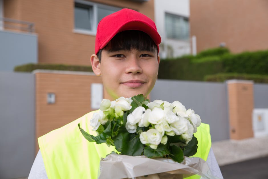 A young male flower delivery courier is standing outdoors in front of modern residential buildings, holding a bouquet of fresh white roses wrapped in white tissue paper and transparent cellophane. He 