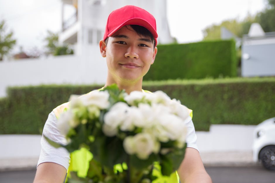 A young delivery person wearing a red cap and a high-visibility vest holding a vibrant bouquet of white flowers with green foliage, standing outdoors in a residential area with greenery and modern hou