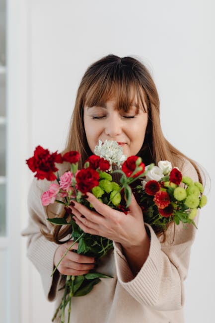 A woman with light brown hair and bangs, dressed in a beige sweater, is holding a vibrant bouquet of fresh flowers close to her face, with her eyes closed and a gentle expression. The bouquet includes