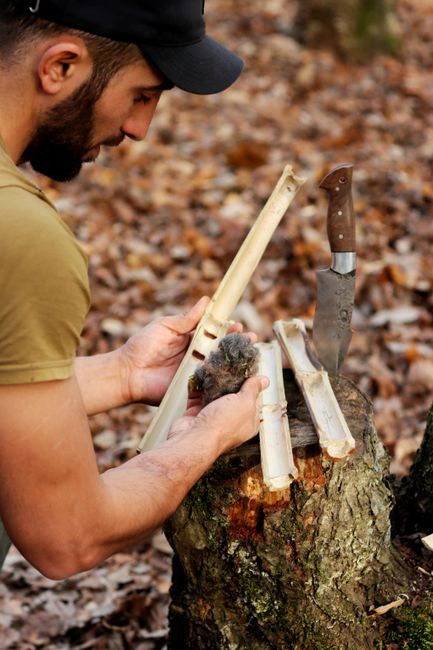 A man with a beard, wearing a black cap and a beige t-shirt, is outdoors in a woodland setting, chopping wood with a fixed-blade knife. He holds a small piece of kindling in one hand and slices it on 