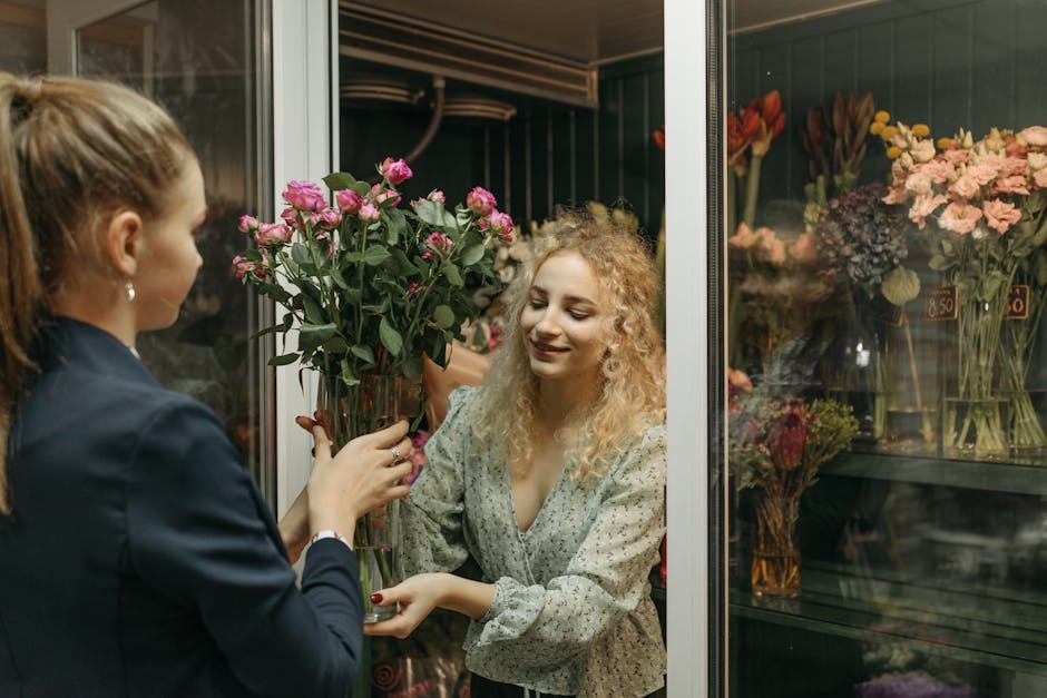 A woman with curly blonde hair, wearing a light-colored dress with small floral patterns, is smiling as she receives a large bouquet of pink roses from a florist. The florist, a woman in a dark blazer