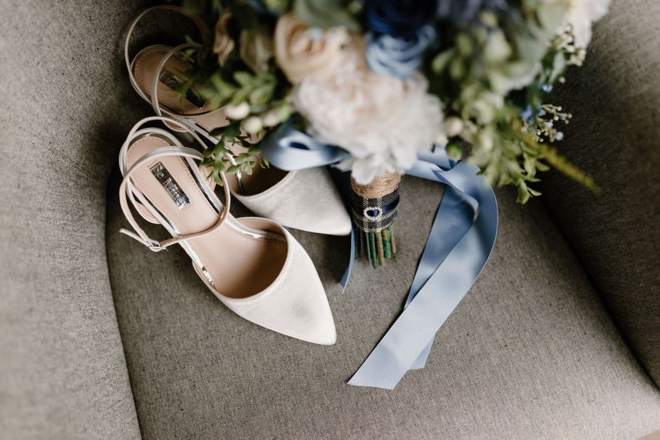 A pair of elegant, cream-colored bridal shoes with pointed toes and ankle straps, resting on a grey fabric surface. Next to the shoes is a bouquet of fresh flowers featuring white roses, blue delphini