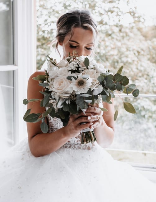 A bride holding a large, elegant bouquet of fresh flowers, including white roses, anemones, and eucalyptus leaves, arranged in a round, lush style. The bouquet is wrapped with a gold ribbon, and the b